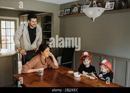 Une famille à une table en bois avec deux enfants habillés comme des pompiers décorant des biscuits avec du chocolat. Banque D'Images