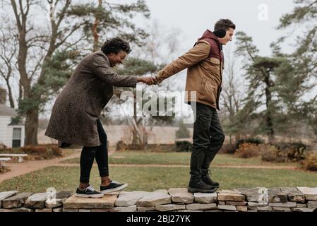 Un homme blanc tenant les mains d'une femme noire lorsqu'ils marchent le long du haut d'un mur dans un jardin. Banque D'Images