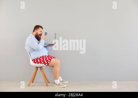 Drôle homme en short rouge avec un ordinateur portable augmente l'information de loupe sur un fond gris. Concept de travailleur indépendant en ligne travaillant à distance. Banque D'Images