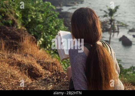 Jolie femme asiatique est assise sur le bord de la montagne avec une vue sur la mer, la lecture d'un livre. À partir de la vue arrière Banque D'Images