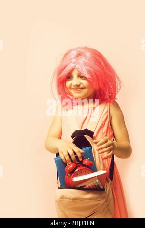 Retour au concept d'école et d'apprentissage. Élève avec visage heureux, isolé sur fond rose clair. Enfant avec réveil et livres. Fille avec perruque dans robe rose avec sac à dos. Banque D'Images