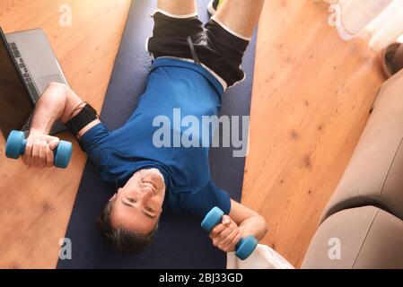 Homme faisant du sport avec des haltères allongé sur le tapis dans le salon à la maison écoutant de la musique avec casque et téléphone. Vue de dessus Banque D'Images