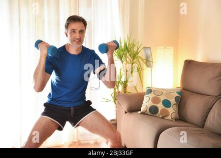 Homme faisant du sport avec des haltères dans le salon à la maison écoutant de la musique avec des écouteurs et le téléphone Banque D'Images
