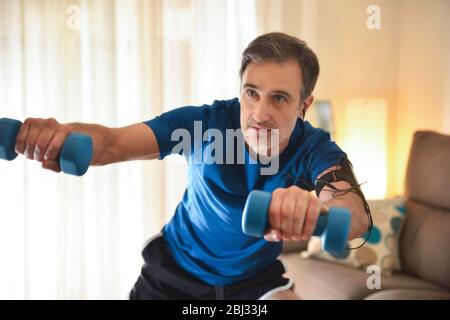 Homme faisant du sport avec des haltères dans le salon à la maison écoutant de la musique avec des écouteurs et le téléphone Banque D'Images