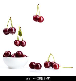 Les cerises rouges mûres avec des bâtons et des feuilles vertes tombent dans un bol en céramique blanc débordent et se dispersent autour d'elle sur un fond blanc isolé. Banque D'Images