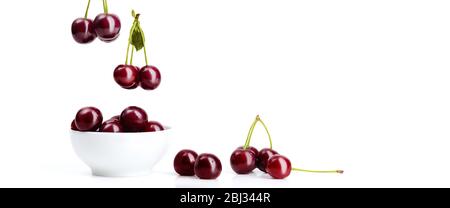 Les cerises rouges mûres avec des bâtons et des feuilles vertes tombent dans un bol en céramique blanc débordent et se dispersent autour d'elle sur un fond blanc isolé. Banque D'Images