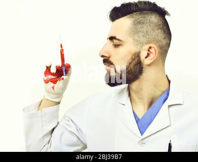 Concept de travail professionnel et de médecine: Médecin avec barbe sombre et expression concentrée du visage regarde la seringue dans sa main, qui est tachée de sang, isolée sur fond blanc Banque D'Images