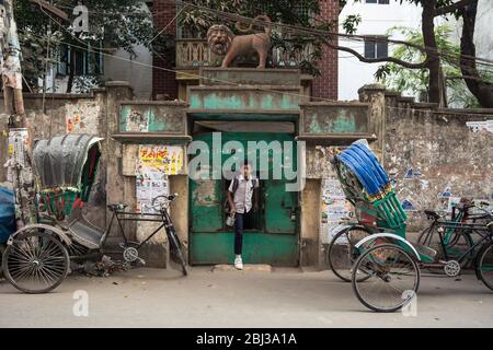 Dhaka / Bangladesh - 14 janvier 2019: Jeune garçon étudiant avec sac à dos sortant de la porte minuscule métallique dans une rue pleine de tuk tuk tuk Banque D'Images