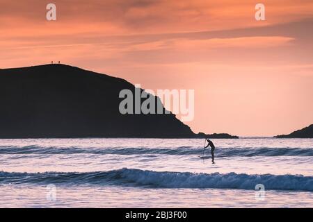 Une paddle-boarder pagayant au-delà de Pentist point East silhouetted par le feu de fin de soirée à Fistral à Newquay, dans Cornwall. Banque D'Images