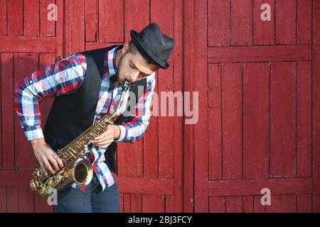 Beau homme heureux dans le chapeau joue de la musique sur le sax devant le fond en bois rouge Banque D'Images