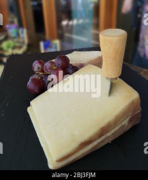 Grande tranche de fromage dur rabouflé avec un instrument de coupe de fromage ou un couteau, sur une assiette en bois associée à des raisins rouges prêts à la dégustation et à l'arrière flou Banque D'Images