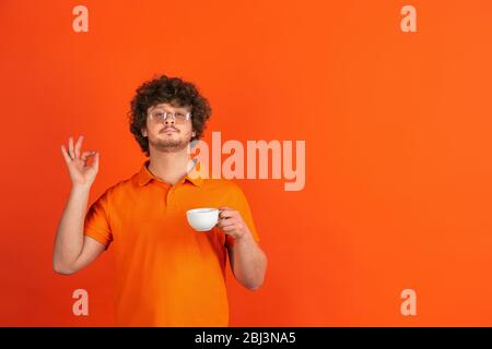 Bon café, enchanté. Portrait monochrome caucasien du jeune homme sur fond de studio orange. Magnifique modèle à plis masculins dans un style décontracté. Concept d'émotions humaines, expression faciale, ventes, publicité. Banque D'Images