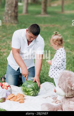 papa et petite fille pour pique-niquer dans le parc le jour de l'été. Le jeune père coupe le pastèque. Le père, le jour de bébé. Des vacances heureux Banque D'Images