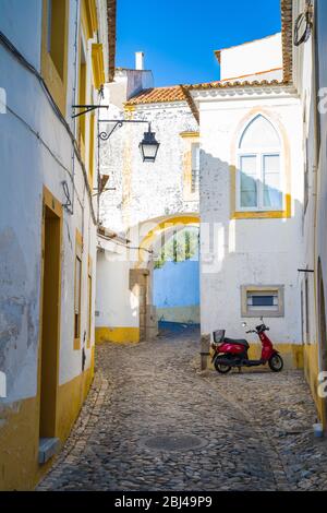 Scooter à moteur stationné dans une rue calme en pierre de galets par des maisons typiques blanches et jaunes à Evora, Portugal Banque D'Images