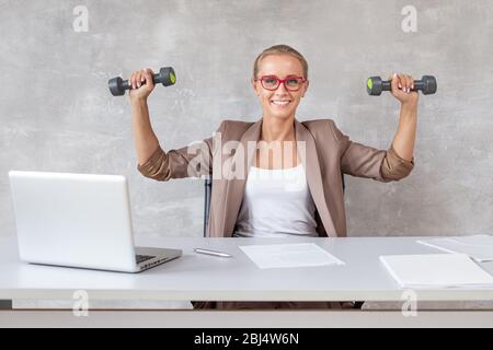 Le travailleur de bureau blond est assis sur une chaise derrière le bureau et tient deux haltères dans les mains. Banque D'Images