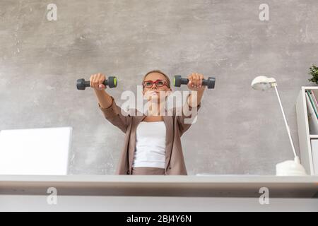 Le travailleur de bureau blond est assis sur une chaise derrière le bureau et tient deux haltères dans les mains. Banque D'Images