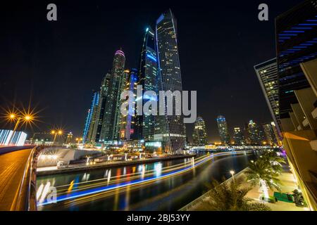 Le sentier lumineux coloré d'un dhow arabe qui traverse la marina et sous ses bâtiments imposants à Dubaï. Banque D'Images