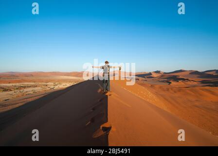 Un seul homme marchant sur une dune de sable avec des armes étirées au coucher du soleil dans le désert de Namide à Sossusvlei, Namibie, Afrique. Format paysage. Banque D'Images