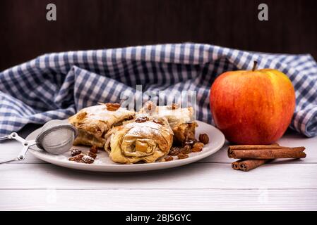 Tranches de strudel à tarte aux pommes maison dans une assiette avec des ingrédients sur une table rustique en bois blanc. Tarte aux pommes, aux raisins secs et à la cannelle, pâtisseries sucrées Banque D'Images