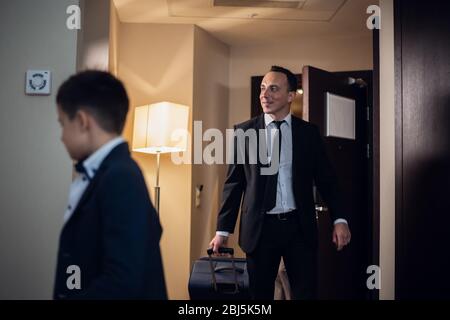 Père et fils en tenue officielle entrant dans une chambre d'hôtel, le père porte une grande valise. Banque D'Images