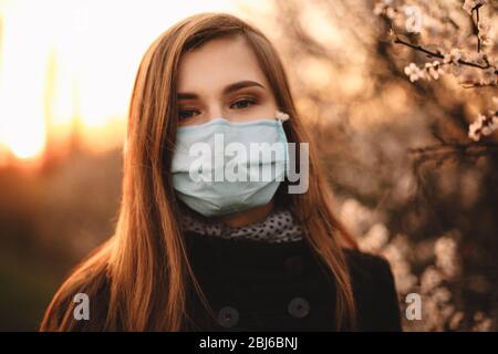 Portrait de la jeune femme fatiguée portant un masque médical de visage debout par arbre fleuri dans le parc au coucher du soleil au printemps Banque D'Images