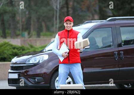 Jeune homme de livraison beau avec boîte et presse-papiers debout près de la voiture, à l'extérieur Banque D'Images