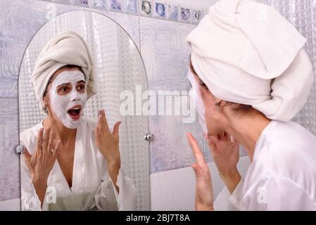 Belle femme brunette à la maison dans la pyjama et serviette sur sa tête mettre le masque blanc sur son visage à la salle de bains Banque D'Images