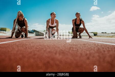 Trois athlètes féminins souriant tout en commençant par les blocs sur la piste de course. Coureurs à des blocs de départ au stade prêt pour la course. Banque D'Images