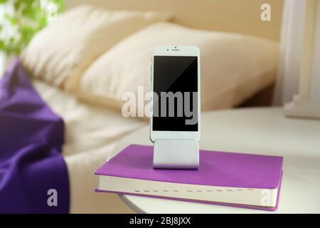 Smartphone avec socle et livre sur une table de chevet dans une chambre Banque D'Images