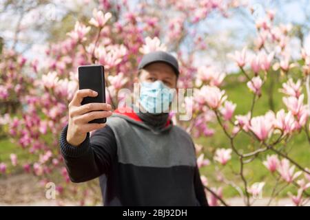 Homme en masque médical faisant selfie avec des fleurs de magnolia pendant la pandémie de COVID-19 Banque D'Images