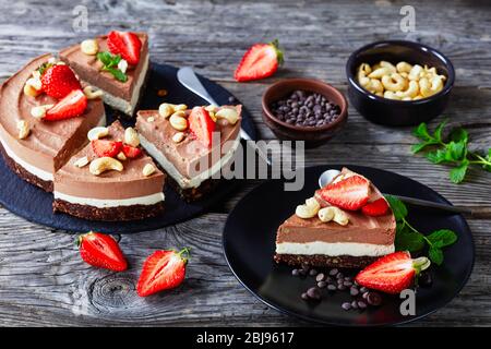 Tarte au triple chocolat cru à la crème de noix de coco et aux noix de cajou trempées avec croûte de dattes brutes, de cacao et de noix sur une plaque noire avec paille Banque D'Images