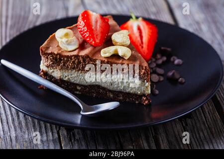 Tarte au chocolat de deux couches de chocolat : blanc et foncé avec croûte non cuite de dattes brutes, cacao et noix sur une plaque noire avec fraises fraîches et c Banque D'Images