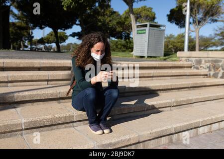 Femme caucasienne portant un masque de protection dans la rue, assise et utilisant son téléphone Banque D'Images