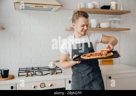 Pizza italienne maison, beau jeune chef mâle dans un tablier sombre tient la plaque de cuisson dans la cuisine lumineuse. Concept de blog vidéo de cuisine Banque D'Images
