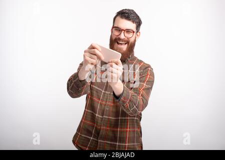 Un homme barbu portant des lunettes est en train de jouer avec son téléphone sur fond blanc. Banque D'Images