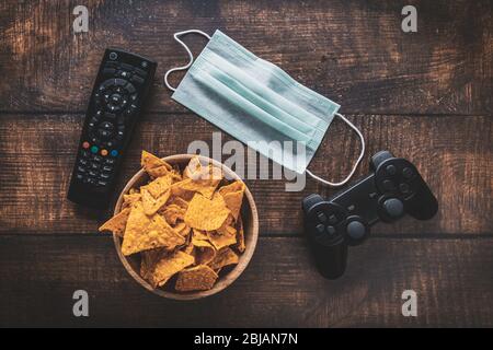 Télécommande, console de jeux rc, bol de chips et masque médical sur table en bois. Coronavirus Covid-19 quarantaine rester à la maison concept. Banque D'Images