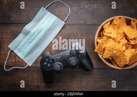 Console de jeu rc, bol de chips et masque médical sur table en bois. Coronavirus Covid-19 quarantaine rester à la maison concept. Banque D'Images