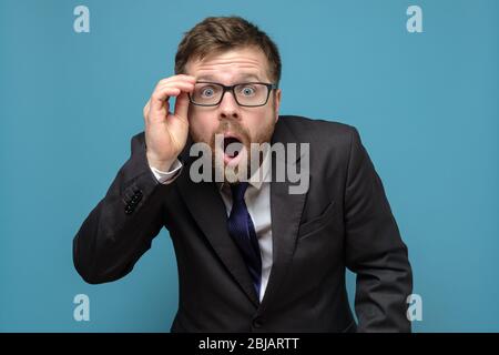 Un homme d'affaires surpris dans un costume tient des lunettes dans la main et en étonnement ouvre la bouche, regarde avec de grands yeux sur l'appareil photo. Banque D'Images