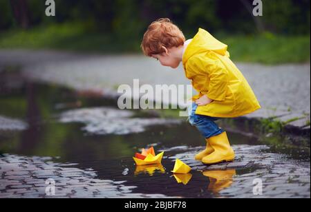 joli petit garçon lance des bateaux en papier dans des flaques de printemps, portant un imperméable et des bottes en caoutchouc Banque D'Images