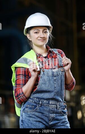 Jeune fille dans une robe de travail et un chapeau blanc dans une usine. Femme dans un uniforme de travail tenant un gilet réfléchissant dans un rangement. Processus de travail. Banque D'Images