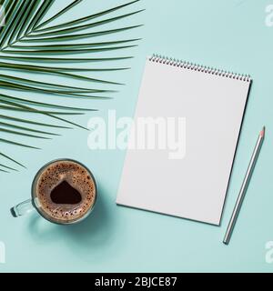 Cahier en papier vierge avec crayon et tasse à café. Livre de croquis en papier vide avec pincil et feuilles tropicales sur fond bleu. Vue de dessus ou plat. Espace de copie pour le texte ou la conception. Banque D'Images