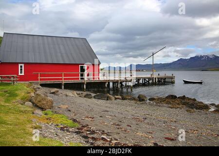 Vue sur le fjord avec jetée, bâtiment traditionnel en bois rouge, bateau de pêche, belles montagnes et îles en arrière-plan, Eskifjordur, Islande Banque D'Images