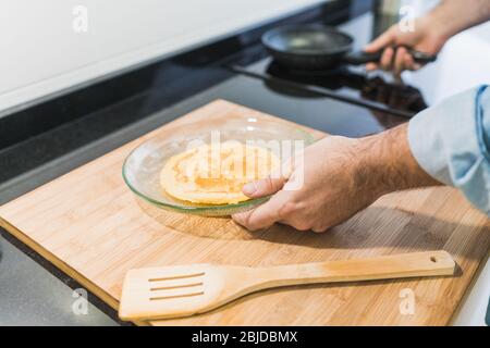 Homme cuisine dans la cuisine dans une chemise en denim. Un homme anonyme tient une poêle avec une crêpe sur une plaque Banque D'Images