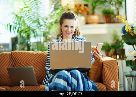 femme de ménage souriante et élégante avec chemisier bleu et veste rayée dans une maison moderne, ensoleillée, avec différents appareils : ordinateur portable, smartphone. Banque D'Images