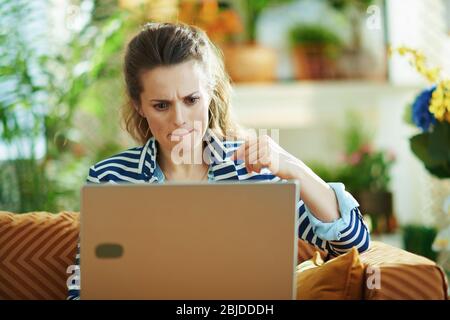 femme à la mode avec chemisier bleu et veste rayée assise sur un canapé avec ordinateur portable chez elle moderne en journée ensoleillée. Banque D'Images