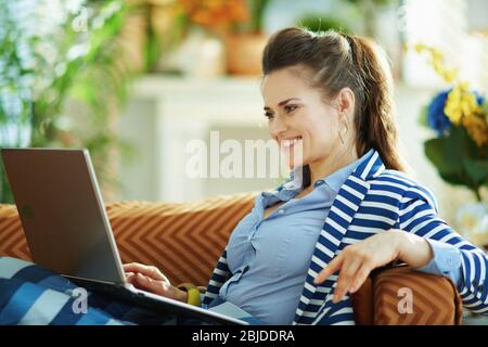 Vue latérale d'une femme heureuse de l'âge moyen avec un chemisier bleu et une veste rayée assise sur un canapé avec ordinateur portable dans le salon moderne en journée ensoleillée. Banque D'Images