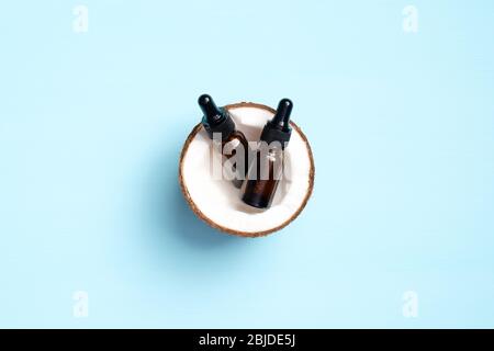 Bouteille d'huile de coco essentielle dans la moitié de la noix de coco sur fond bleu. Plat, vue du dessus. SPA cosmétique biologique naturelle, soins de la peau, soins du corps conceps Banque D'Images