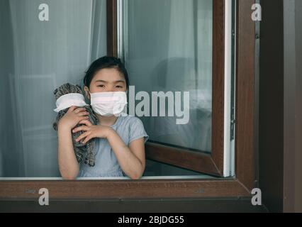 Une fille dans un masque avec un jouet farci se tient à la fenêtre ouverte de sa maison Banque D'Images
