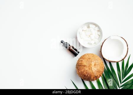 Bouteille d'huile de coco essentielle avec noix de coco tranché dans un bol, feuille de palmier tropicale, moitié de noix de coco sur fond blanc. Plat, vue du dessus. ORG. NATURELLE DU SPA Banque D'Images