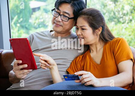 Un couple asiatique expérimenté et heureux regarde leur tablette avec une carte de crédit prête à payer au salon dans leur maison Banque D'Images